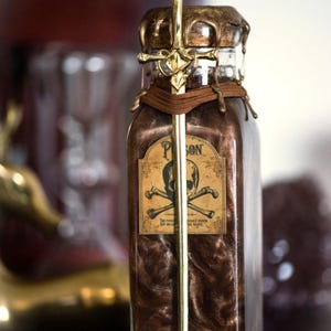 Peut inclure: Une bouteille en verre avec un couvercle doré et une épée qui sort du haut. La bouteille est remplie d'un liquide sombre et porte une étiquette indiquant "Poison" avec un crâne et des os croisés. Le texte "Lune Rouge Mystique" est écrit sous la bouteille.