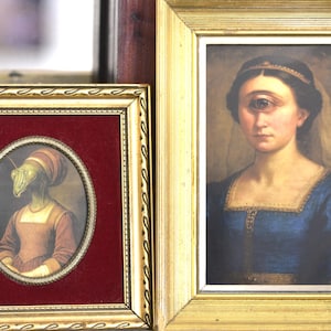 Puede incluir: Dos obras de arte enmarcadas. Una presenta un retrato de una mujer con un solo ojo, vestida con un vestido azul y una diadema dorada. La otra es un retrato ovalado de un insectoide en un marco rojo y dorado.