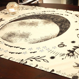 May include: A beige tapestry with a black and white illustration of a crescent moon, stars, and floral designs. The tapestry is laid out on a wooden table.