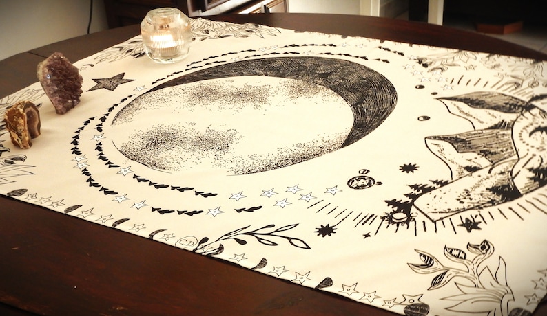 May include: A beige tapestry with a black and white illustration of a crescent moon, stars, and floral designs. The tapestry is laid out on a wooden table.