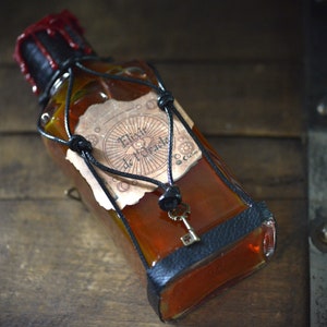 Può includere: Una bottiglia di vetro con un sigillo di cera rossa e un cinturino in pelle nera. La bottiglia contiene un liquido arancione scuro e ha un'etichetta che recita "Fleur de Miracle".