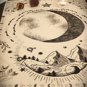 May include: A beige cloth tapestry with a black and white illustration of a crescent moon, stars, mountains, and trees. The tapestry has a bohemian aesthetic.