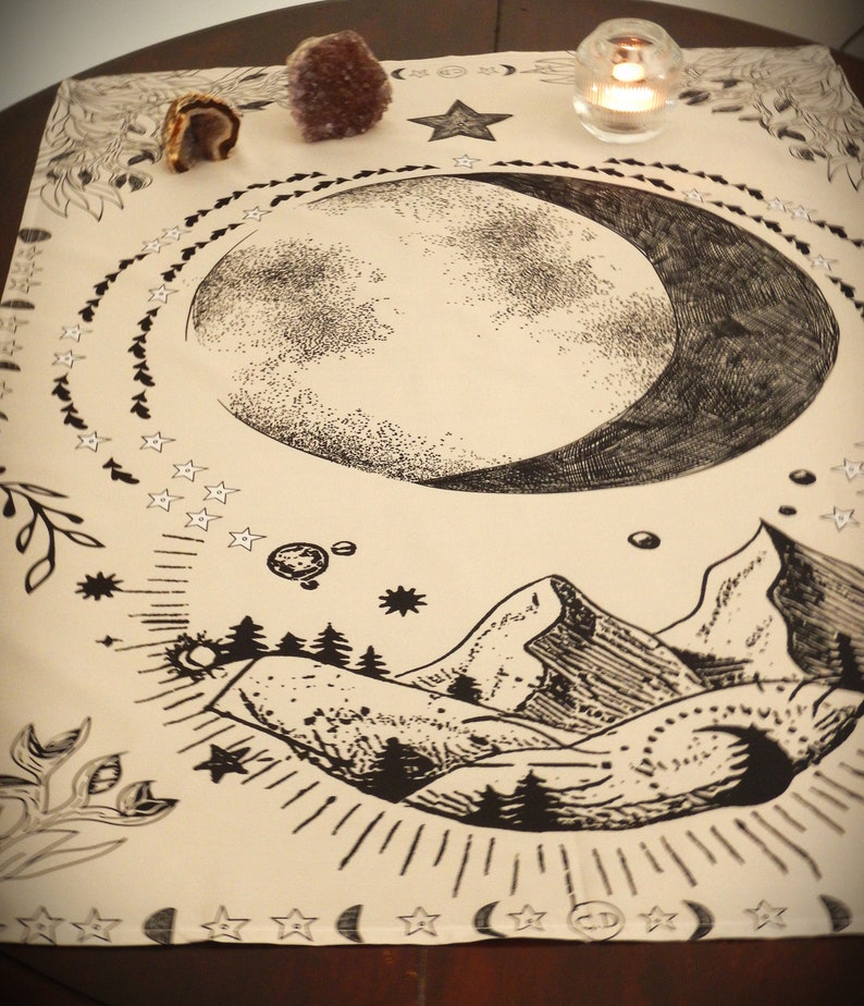 May include: A beige cloth tapestry with a black and white illustration of a crescent moon, stars, mountains, and trees. The tapestry has a bohemian aesthetic.