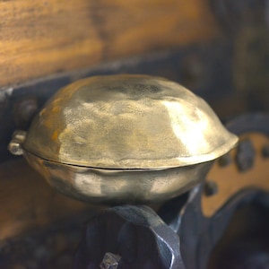 Könnte beinhalten: Ein messingfarbener Behälter mit Deckel und runder Form. Der Behälter hat eine strukturierte Oberfläche und einen Scharnierverschluss. Der Hintergrund zeigt Holz- und Metallelemente, die eine Vintage- oder antike Ästhetik suggerieren.