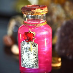 Op de afbeelding: Een roze drankfles met een gouden ketting en een rode hartvormige hanger. De fles heeft een label met "Love Potion" en is verzegeld met een bruine wasdop. De drank binnenin heeft een glinsterend effect.