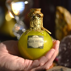Könnte beinhalten: Eine kleine, runde, grüne Glasflasche mit goldenem Verschluss und einem Etikett mit der Aufschrift "Cresent Apothecary Aging Potion".