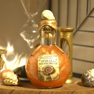 Op de afbeelding: Een decoratieve drankfles met een sleutelvormige stopper en een label met de tekst "SPIRALIS MEMORIAE". De fles is gevuld met een glinsterende oranje vloeistof en bekroond met een zeeschelp. De fles staat op een houten oppervlak.