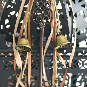 Può includere: Lanterna in metallo nero con intagli decorativi, ornata da una chiave in bronzo, piccole campane in ottone e nastri beige. La lanterna è sospesa a una corda di iuta, creando un'estetica vintage.
