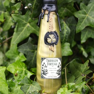 Peut inclure: Une bouteille décorative en verre avec un sceau de cire noire et un emblème de trèfle à quatre feuilles. La bouteille contient un liquide jaune et porte une étiquette "Trifolium Fortunae". Entourée de lierre vert.