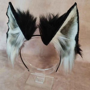 May include: A pair of black and white faux fur animal ears attached to a black headband. The ears are triangular with black fur on the outside and white and gray fur on the inside. The headband is displayed on a clear stand.