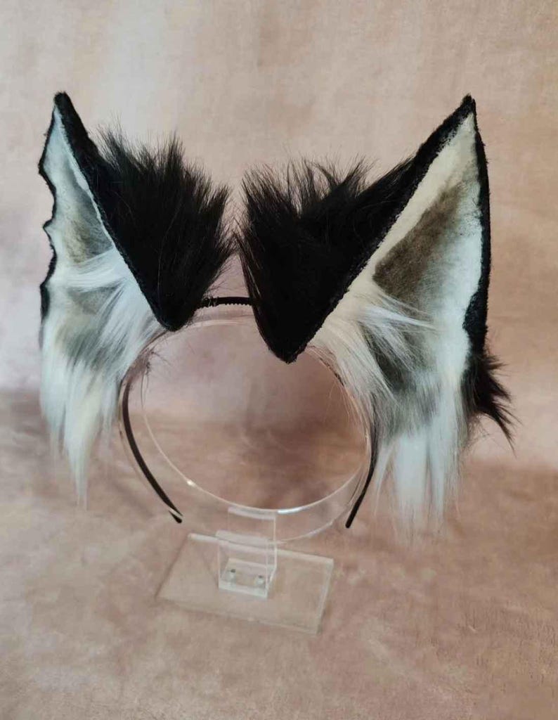 May include: A pair of black and white faux fur animal ears attached to a black headband. The ears are triangular with black fur on the outside and white and gray fur on the inside. The headband is displayed on a clear stand.