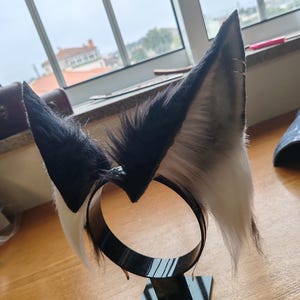 May include: A black and white faux fur headband with upright, pointed ears. The ears are black on the outside, transitioning to gray and white on the inside. The headband is black and sits on a black stand.