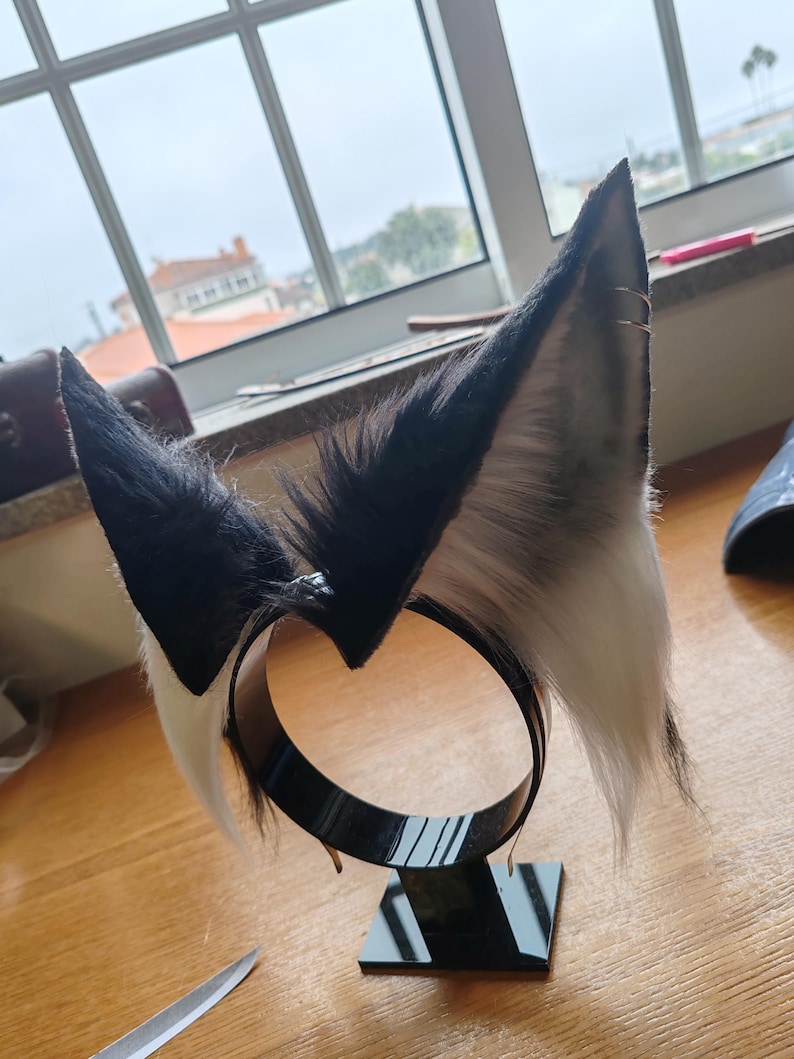 May include: A black and white faux fur headband with upright, pointed ears. The ears are black on the outside, transitioning to gray and white on the inside. The headband is black and sits on a black stand.