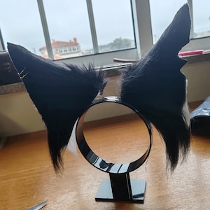 May include: Black faux fur cat ears attached to a black headband. The ears are upright and pointed, with a fluffy texture. The headband is circular and sits on a small black base. A pair of scissors is visible on the wooden surface.