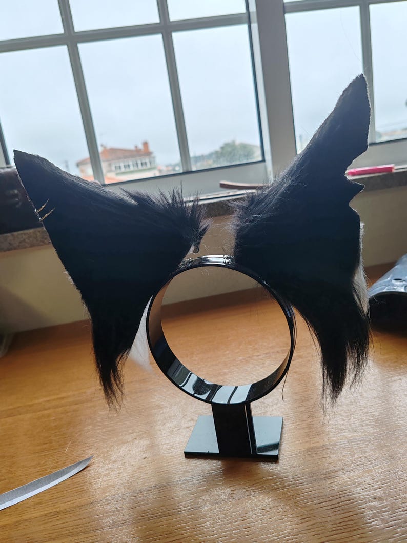 May include: Black faux fur cat ears attached to a black headband. The ears are upright and pointed, with a fluffy texture. The headband is circular and sits on a small black base. A pair of scissors is visible on the wooden surface.