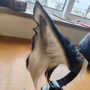 May include: A pair of black and white faux fur wolf ears attached to a black metal headband. The ears have a pointed shape with a dark gray interior and a white fur edge. The headband is on a black square base.