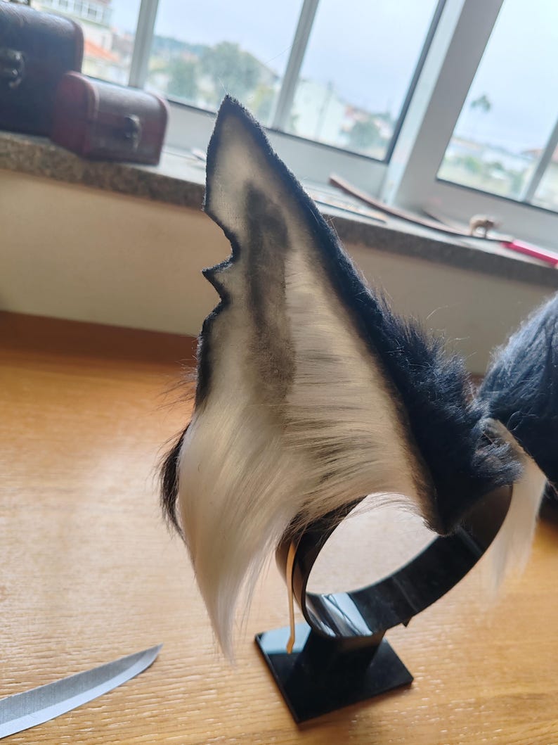 May include: A pair of black and white faux fur wolf ears attached to a black metal headband. The ears have a pointed shape with a dark gray interior and a white fur edge. The headband is on a black square base.