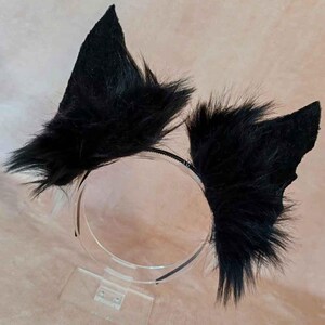May include: Black faux fur cat ears attached to a thin black headband. The ears are triangular and fluffy, with a pointed shape. The headband is displayed on a clear acrylic stand. The overall aesthetic is playful and costume-like.