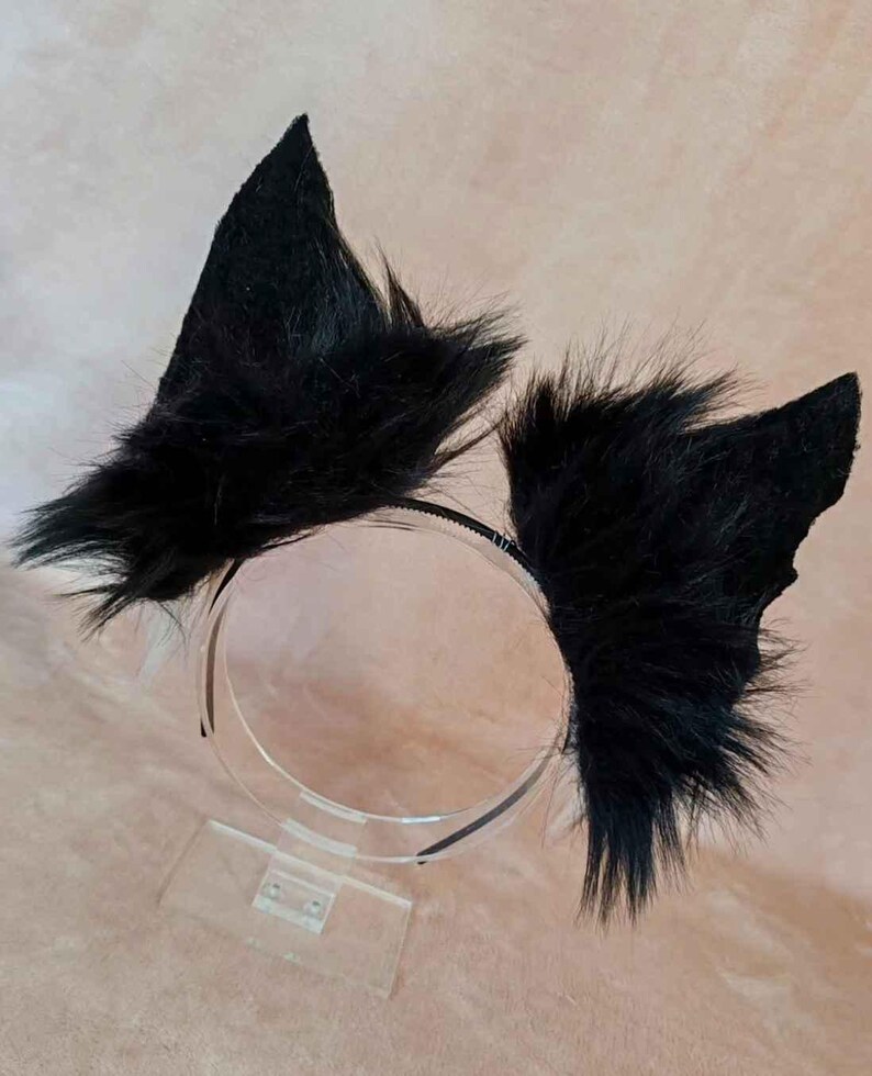 May include: Black faux fur cat ears attached to a thin black headband. The ears are triangular and fluffy, with a pointed shape. The headband is displayed on a clear acrylic stand. The overall aesthetic is playful and costume-like.