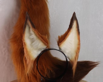 May include: A set of fox ears and tail, featuring reddish-brown faux fur. The ears are attached to a black headband, with a cream-colored interior and dark brown tips. The fluffy tail is also reddish-brown.