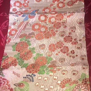 May include: A detailed textile with a floral and bird pattern. The design features red and orange flowers, green leaves, and a bird with blue, red, and white feathers. The background is a light beige color with heart-shaped patterns.