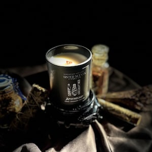 Puede incluir: Una vela de boticario negra en un tarro de cristal con el texto "Sister Scents Candle Co." y "Apothecary". La vela está sobre un soporte negro decorativo. La vela tiene una mecha de madera y está rodeada de otros artículos de boticario.