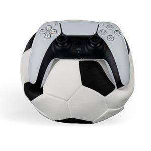 May include: A white and black soccer ball with a white and black PlayStation 5 controller resting on top. The controller has black accents and the PlayStation symbols.