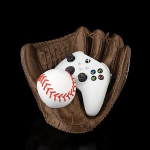 Op de afbeelding: Een bruine honkbalhandschoen houdt een witte honkbal met rode stiksels en een witte Xbox-controller vast. De controller heeft zwarte knoppen en gekleurde actieknoppen. De achtergrond is zwart.