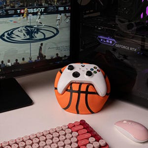 May include: A white Xbox controller sits on an orange basketball-shaped stand. A pink and red keyboard and a pink mouse are in the foreground. A computer monitor shows a basketball game. A computer tower is in the background.