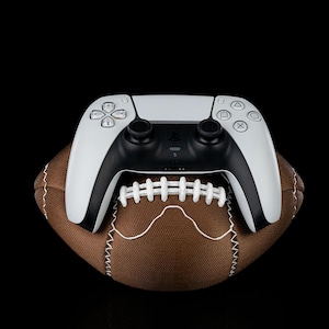 Puede incluir: Un mando de videojuegos blanco y negro descansa sobre un balón de fútbol americano marrón. El mando tiene un diseño elegante con botones y joysticks. El balón tiene costuras blancas y una forma clásica, sobre un fondo negro.