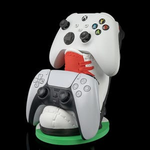 May include: A white and black gaming controller stand with a green base. The stand holds two game controllers, one Xbox and one PlayStation. The Xbox controller is white with colored buttons. The PlayStation controller is white and black.