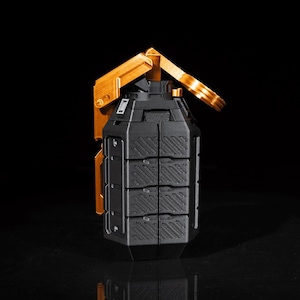 May include: A black and gold grenade-shaped object with a textured surface. The gold-coloured handle is raised, and the body is segmented. The object is set against a black background.