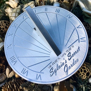 May include: A silver metal sundial with Roman numeral markings and a decorative inscription that reads "Sylvia's Secret Garden" and includes the latitude and longitude coordinates.