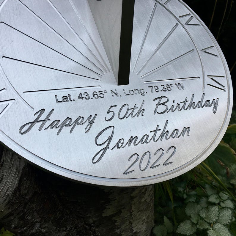 May include: A silver-colored sundial with the inscription "Happy 50th Birthday Jonathan 2022" and coordinates "Lat. 43.65&deg; N, Long. 79.38&deg; W".