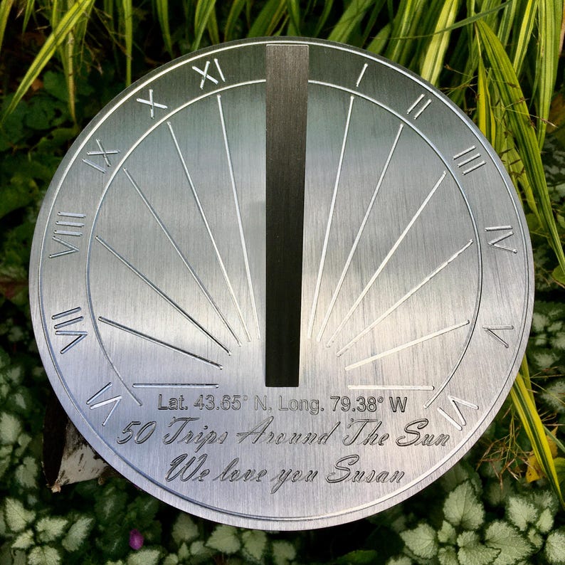 May include: A silver metal sundial with Roman numerals and a black gnomon. The sundial is engraved with the text "Lat. 43.65&deg; N. Long. 79.38&deg; W 50 Trips Around The Sun We love you Susan".