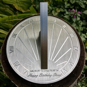 May include: A silver metal sundial with Roman numerals and a gnomon. The sundial is engraved with the text "Lat. 48.85&deg; N, Long. 79.38&deg; W" and "Happy Birthday Steve!"