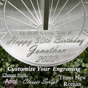 May include: A silver-colored sundial with a personalized inscription that reads "Happy 50th Birthday Jonathan 2022". The sundial also includes the coordinates "Let. 43.65&deg; N, Long. 79.38&deg; W".