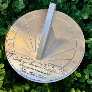 May include: A silver metal sundial with Roman numerals and a gnomon. The sundial is engraved with the text "Emily and Connor's First Home Loving memories since Feb. 23rd, 2023" and the coordinates "Lat. 43.71° N, Long. 79.35° W".
