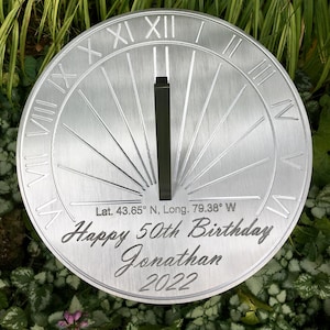 May include: A silver metal sundial with Roman numerals and a black gnomon. The sundial is engraved with the text "Lat. 43.65&deg; N, Long. 79.38&deg; W Happy 50th Birthday Jonathan 2022".