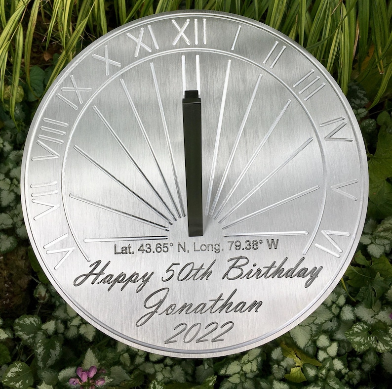 May include: A silver metal sundial with Roman numerals and a black gnomon. The sundial is engraved with the text "Lat. 43.65&deg; N, Long. 79.38&deg; W Happy 50th Birthday Jonathan 2022".