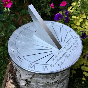 Könnte beinhalten: Eine silberne Sonnenuhr aus Metall mit römischen Ziffern und einem Gnomon. Die Sonnenuhr hat eingravierten Text, einschließlich Koordinaten und Datum. Die Sonnenuhr steht auf einem Birkenstamm, umgeben von Blumen.