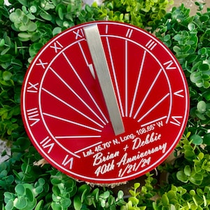May include: Red metal sundial with white Roman numerals and a silver metal gnomon. The sundial is personalized with the text "Lat. 45.70° N, Long. 108.65° W Brian + Debbie 40th Anniversary 1/21/24".