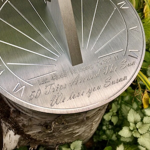 May include: A silver metal sundial with the inscription "Lat. 43.65&deg; N. Long. 79.38&deg; W 50 Trips Around The Sun We love you Susan".