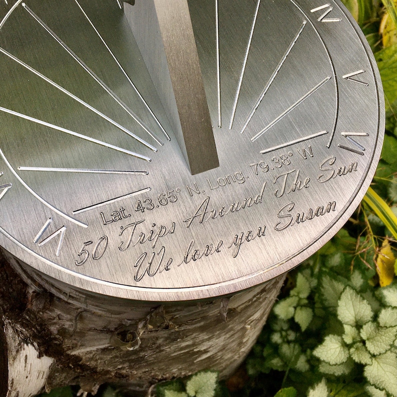 May include: A silver metal sundial with the inscription "Lat. 43.65&deg; N. Long. 79.38&deg; W 50 Trips Around The Sun We love you Susan".