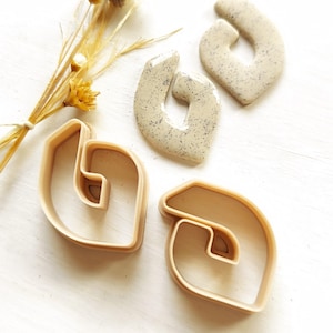 Puede incluir: Dos pares de pendientes de arcilla polimérica beige en forma de línea curva. Los pendientes están moteados de blanco y tienen un acabado mate. Los pendientes se muestran junto a dos cortadores de galletas de plástico beige de la misma forma.