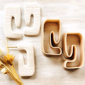 May include: Two pairs of off-white ceramic earrings with a unique abstract shape. The earrings are shown next to a pair of silicone molds for making the earrings.