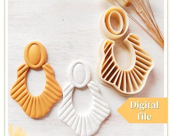 Pode incluir: Uma vista superior mostra dois brincos de argila acabados e um conjunto de cortadores de brincos. Um brinco é amarelo mostarda, o outro é branco. Os cortadores são bege. Os brincos têm uma parte superior oval e uma parte inferior em forma de leque. O texto numa caixa amarela diz "Digital file."