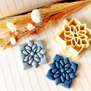 May include: A light blue and a dark blue textured flower-shaped clay piece alongside a cream-colored plastic flower-shaped cookie cutter. The items are arranged on a lined surface with dried white and tan flowers positioned at the top.