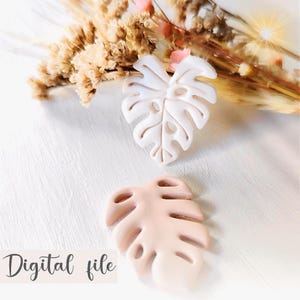Puede incluir: Dos adornos decorativos con forma de hoja de monstera en blanco roto y melocotón. Las hojas están sobre una superficie blanca con flores secas en el fondo. El texto "Digital file" está en la parte inferior izquierda.