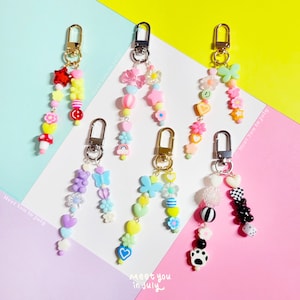 May include: Five colorful beaded keychains with different charms, including hearts, stars, butterflies, and flowers. The keychains are attached to gold-colored key rings. The text "Meet you in July" is written on the pink background.